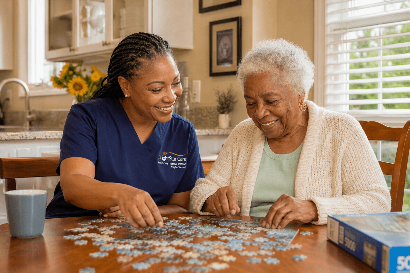 A warm, professional in-home caregiver sits with an elderly woman doing a puzzle at a kitchen table while sunlight streams through the window, both smiling and engaged. Diverse Georgia home setting, gentle and reassuring mood, soft natural daylight, photorealism, professional editorial photography, warm tones, 3:2 aspect ratio