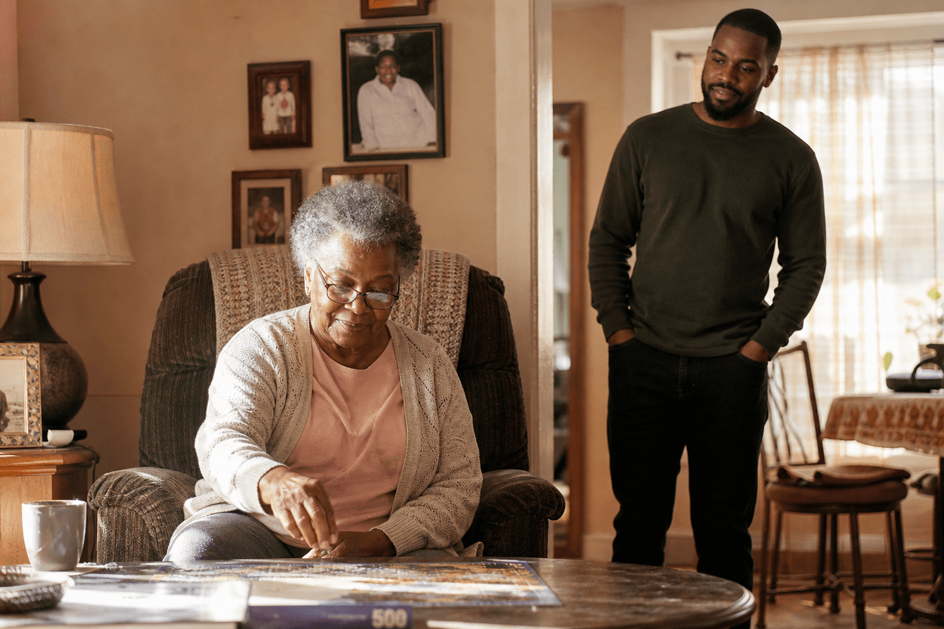 An adult son quietly observes his elderly mother in her home, noticing details around her, a look of gentle concern on his face as he takes in how she is managing day to day. Diverse Georgia family, thoughtful and caring mood, warm afternoon light in a comfortable lived-in living room, photorealism, professional editorial photography, warm tones, 3:2 aspect ratio